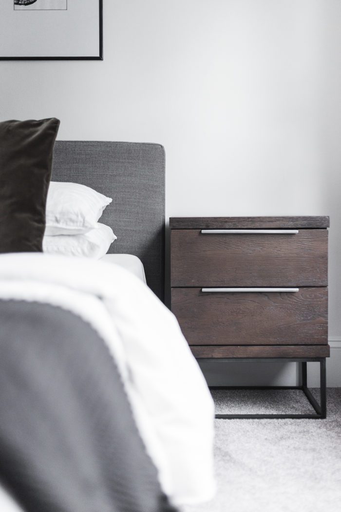 Dark wood nightstand next to gray headboard with white bedding. 