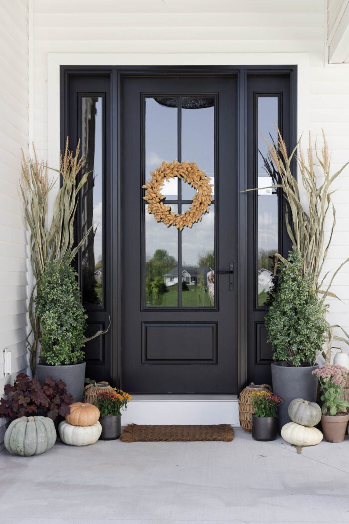Black front door decorated for Fall with plants, a wreath, and corn stalks. 